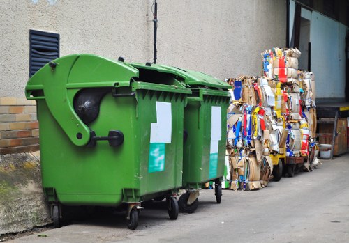 A row of three large, closed, green wheelie bins positioned on a paved driveway or street beside a building with a beige, textured exterior wall and a small black ventilation grille. The bins are made of durable plastic with smooth, matte finishes, featuring white labels or stickers on their front panels. To the right of the bins, there are multiple compacted bales of waste materials, including cardboard and paper, bound with plastic straps and stacked against the building wall. The surrounding environment appears to be an urban or suburban area, possibly in or near Manor Park, with the bins used for rubbish collection services. The scene is captured in natural daylight, emphasizing the contrast between the vibrant green bins and the neutral background of the wall and paved surface. This setup aligns with waste management practices offered by a company like Commercial Waste Removal Manor Park for domestic or commercial refuse disposal.