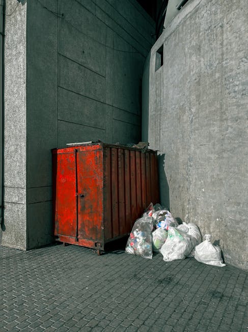 An outdoor scene showing scattered discarded cardboard boxes, some flattened and others partially intact, amidst patches of grass and dirt along a sloped area near a fence. The boxes are mainly green 