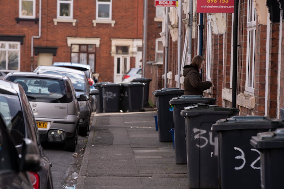 A row of dark grey wheelie bins with white numbering along a residential pavement, positioned in front of brick terraced houses with white-framed windows and stone lintels. A woman dressed in a brown 